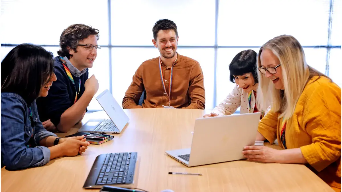 A group of four colleagues sit around a laptop laughing