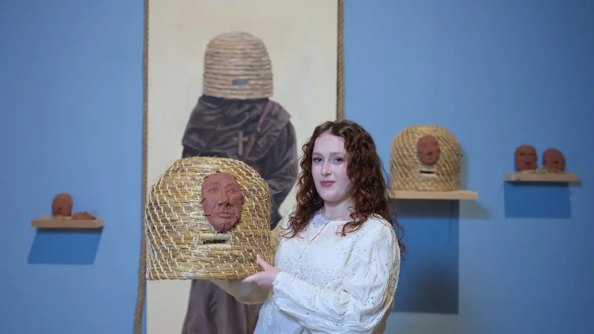 An artist stands in front of her exhibition and holds a piece featuring a woven basket with clay faces.