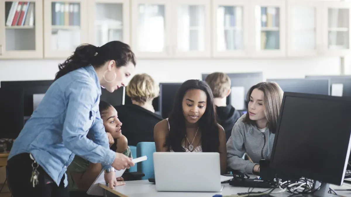 Mature teacher assisting female students using laptop in computer lab at high school - stock photo