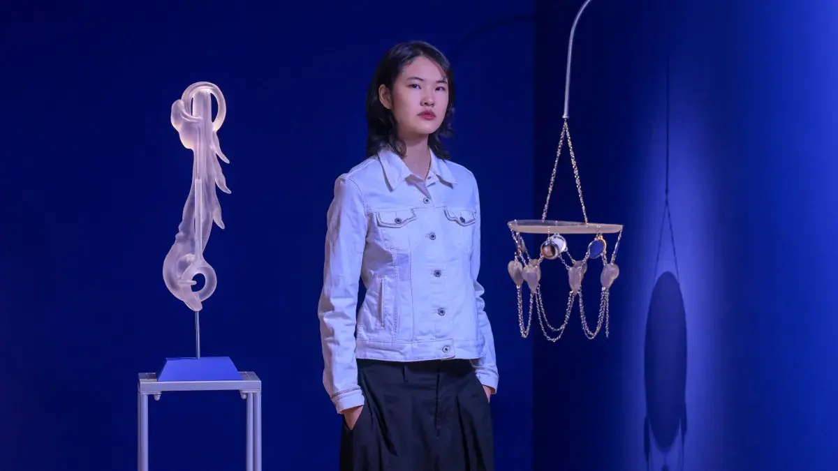 An artist stands in front of their exhibition and holds a piece featuring a woven basket with clay faces.