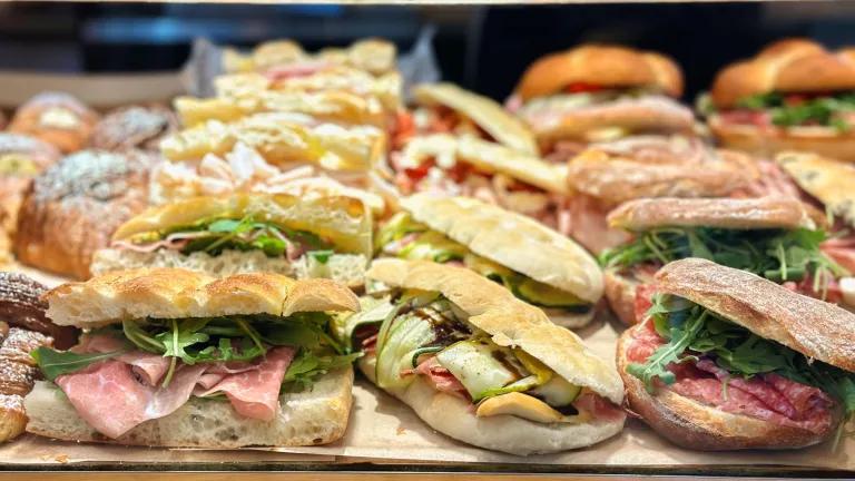 Close-up of multiple rustic sandwiches displayed on a bakery counter, featuring crusty bread filled with a mix of cured meats and salad