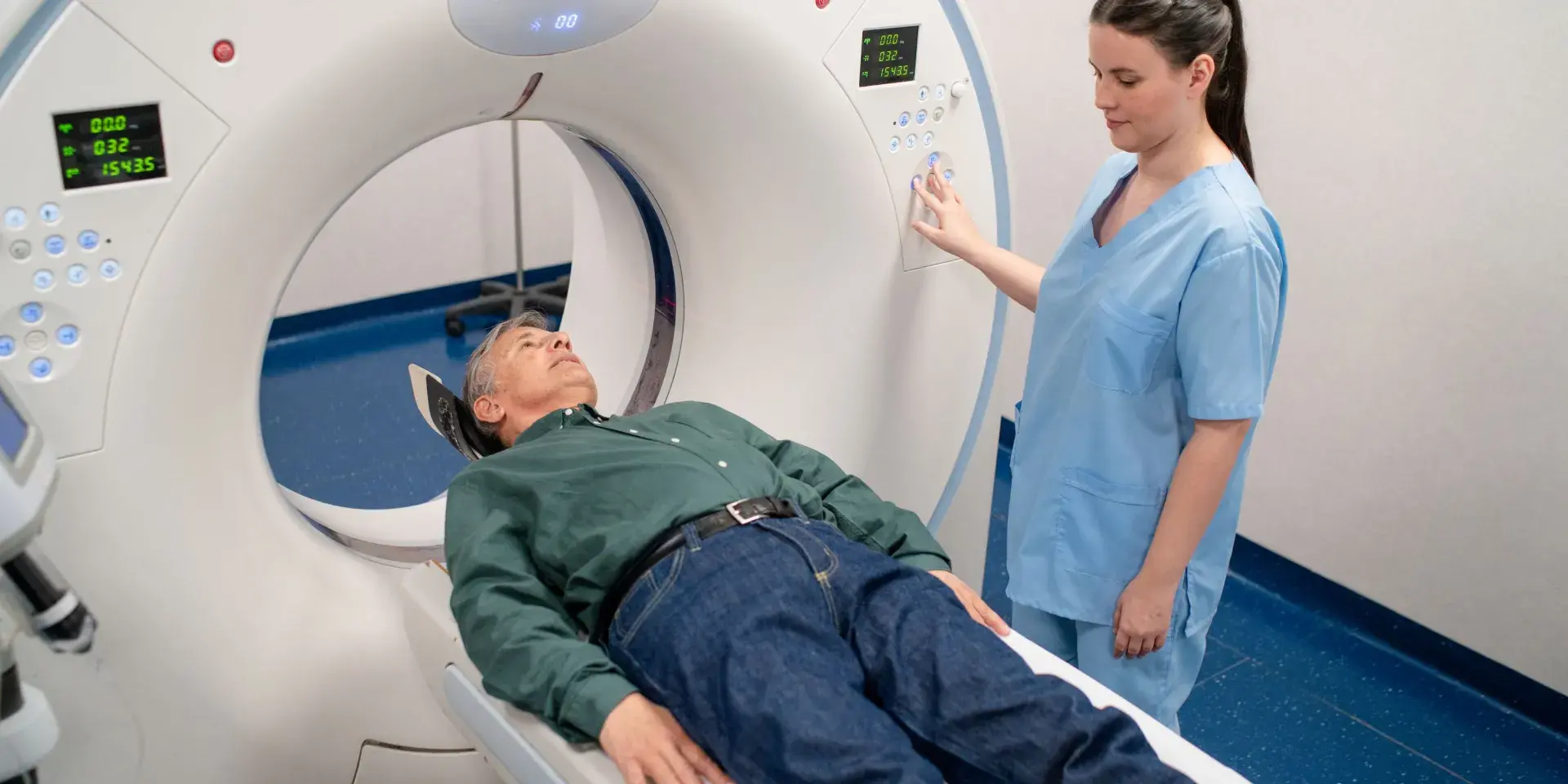 Older male patient on a CT scan machine in a hospital with a nurse in blue scrubs operating the equipment