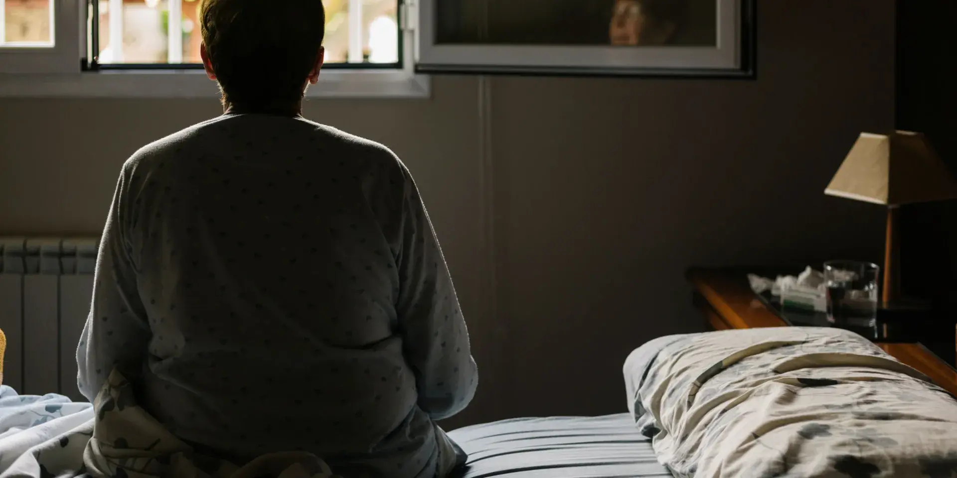 Older woman pictured from behind, sat on a bed looking out of a window