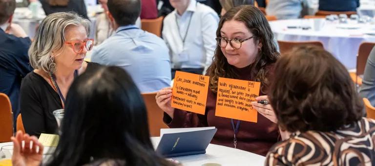 A woman wearing glasses sitting at a table holds up two flash cards to three other people at table outlining details of a discussion that they have just had about AI infrastructure. 