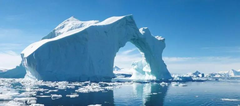 A large iceberg with pieces of ice floating in the ocean in the foreground and blue sky in the background