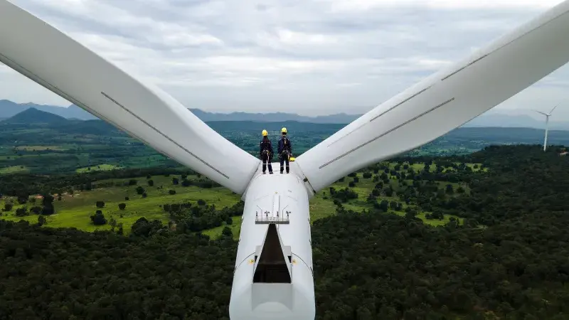 Two people in yellow hard hats stand on top a wind turbine and look at green countryside