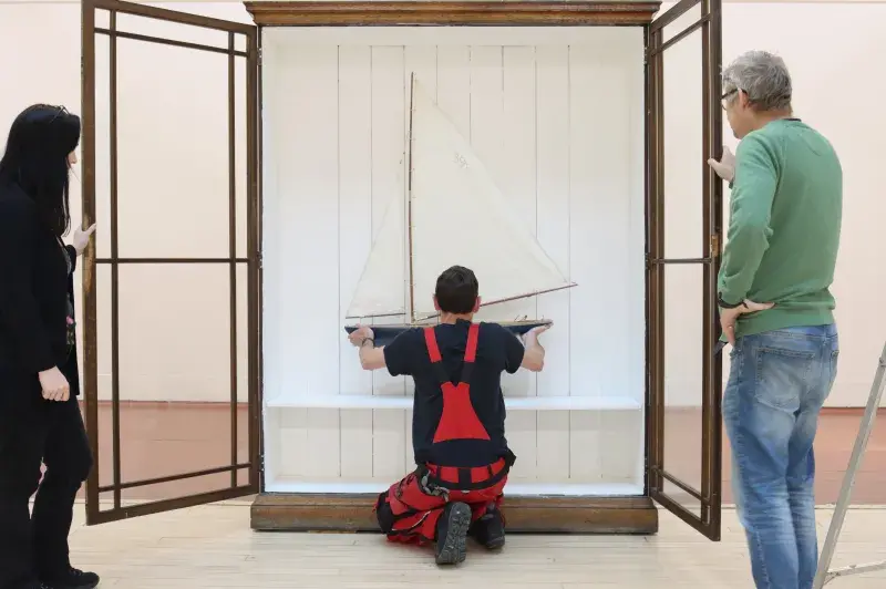 Two people watch the instalation of a model boat against white wooden paneling inside a dark wooden case with glass windows in the doors.