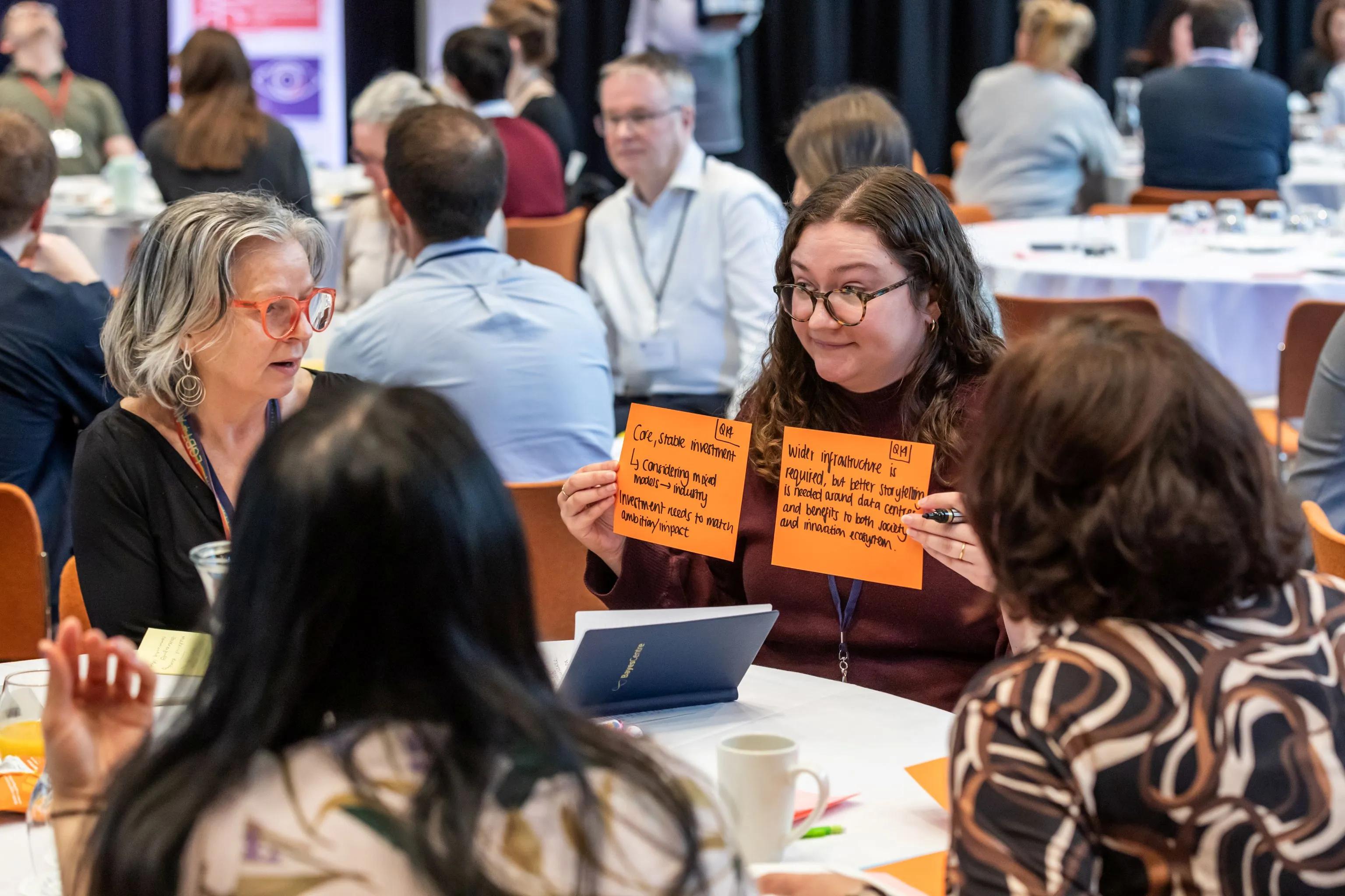 A woman wearing glasses sitting at a table holds up two flash cards to three other people at table outlining details of a discussion that they have just had about AI infrastructure. 
